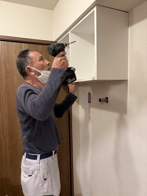 練馬区のH様邸では家具屋による吊戸の設置が行われていました。 74㎝の洗濯機パンを使うので、上部に75㎝幅のオープン棚収納を設けました。 簡単なオープン棚や既製品でも良さそうな場所にわざわざ造作家具を使ったのには理由があります。 もともとこちらのお宅は洗濯機の上にメッシュのオープン棚が1枚あり、そこにフェイスタオルを平置きしてたくさん積んでお使いでした。 というのも、H様は手を洗ったら都度新しいタオルを使う習慣があり（ですのでタオル掛けは必要ありませんでした）、タオルを沢山お使い、かつすぐに手に取れるようオープンな状態が良かったのです。 棚板ではタオルを積んだ時にずれて雑多に見えがちなこと、そしてそれまでは足元に置いて使われていた洗剤も置けるように、ハコ型が良いと思いました。 さらに梁下ということで寸法の制限もあり、わざわざ造作にしたわけです。 前もって伝えておいた形で造ってきてくれ、現場では取付けのみ。 あっという間に取付け完了、ついでと言ってはなんですが、隣のトイレの吊戸棚（こちらは既製品）も取付けしてもらいました。 ささやかな場所のささやかな工夫ですが、生活の中で満足感に大きな差があるのでは…と思っています。